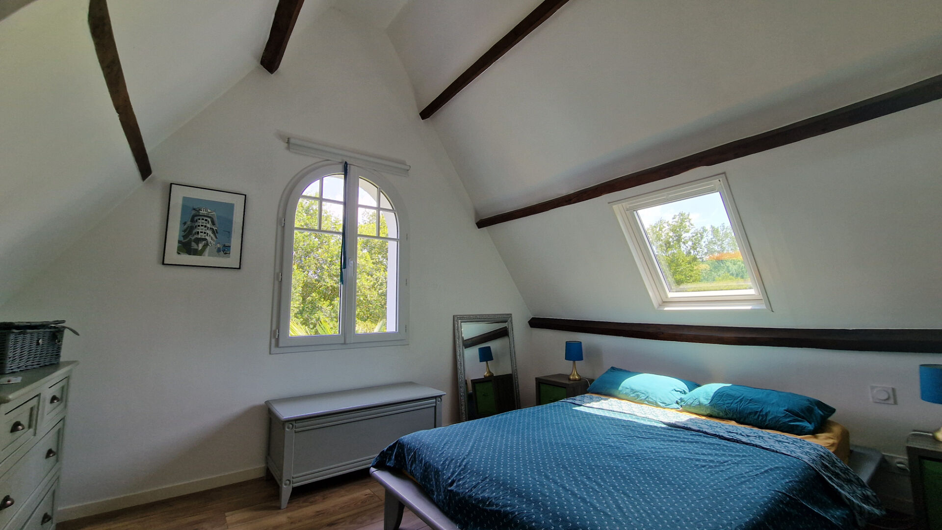 Bright bedroom with sloped ceiling and beams, featuring a double bed and window overlooking greenery in Côtes-d'Armor